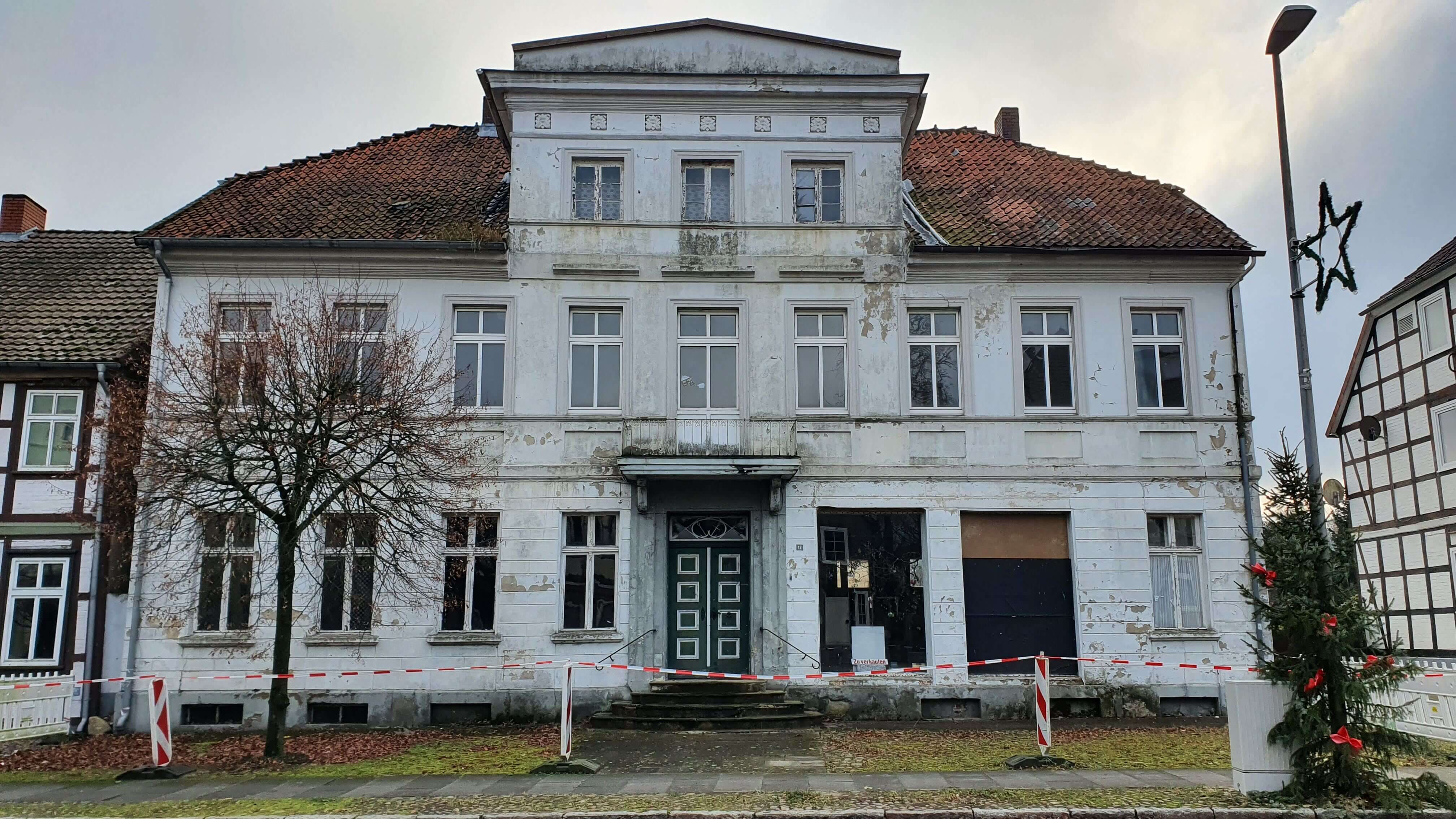 Ein altes, zweistöckiges Gebäude mit abblätternder Fassade und einem vernachlässigten Erscheinungsbild. Einige Fenster sind mit Brettern vernagelt. Vor dem Gebäude sind Absperrbänder gespannt, die den Zutritt verhindern. Links steht ein kahler Baum, rechts ist ein Teil eines Weihnachtsbaumes mit roten Kugeln zu sehen. Der Himmel ist bewölkt.