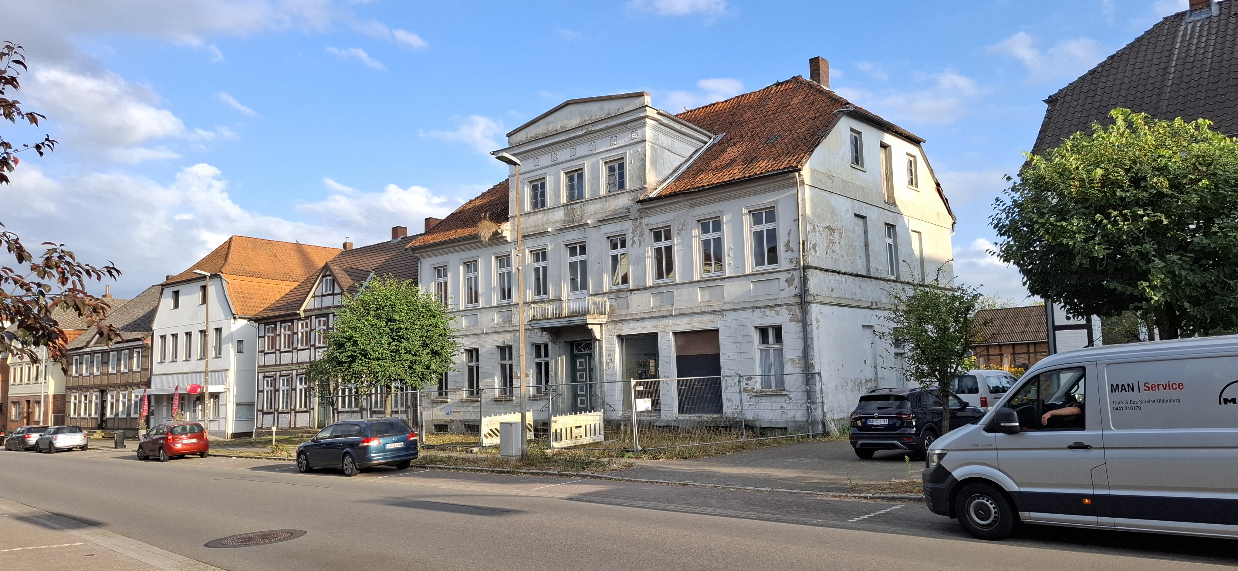 Straßenansicht mit einem großen, alten und sanierungsbedürftigen Wohnhaus mit bröckelnder Fassade zwischen weiteren historischen Häusern, davor parkende Autos und ein Lieferwagen.